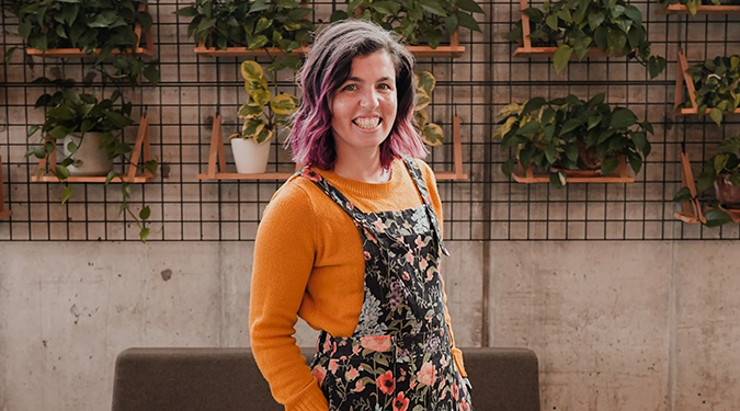 Woman with purple-tinted hair smiles warmly, wears a floral dress over an orange sweater. Behind her, potted plants hang on a wire wall, adding greenery.