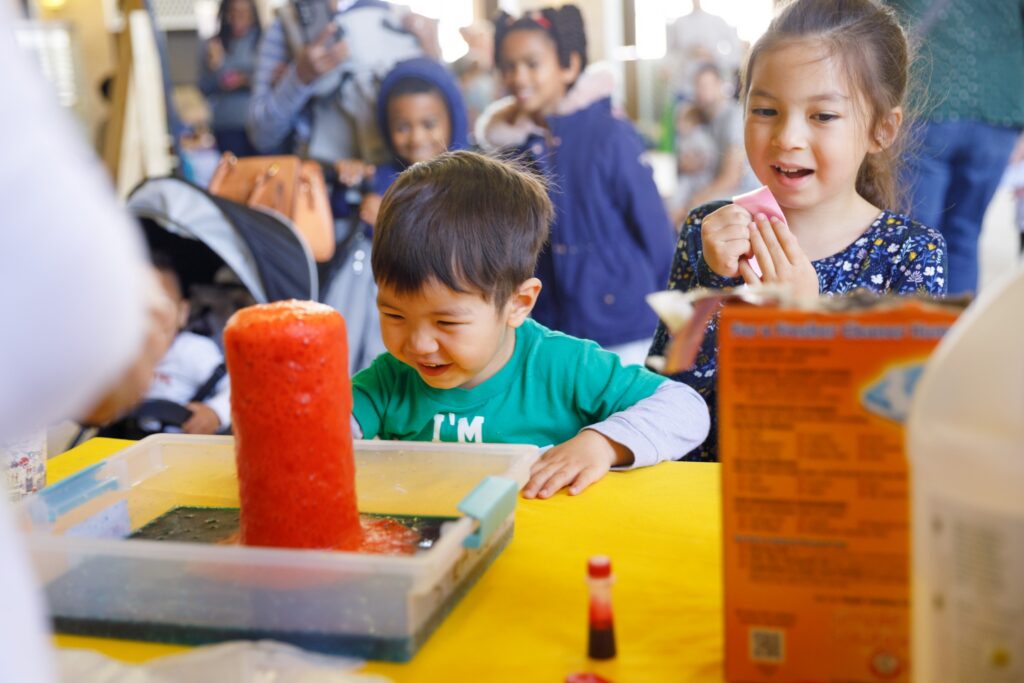 Two children excitedly watch a red baking soda volcano erupt on a yellow table, surrounded by onlookers in a lively science fair setting.