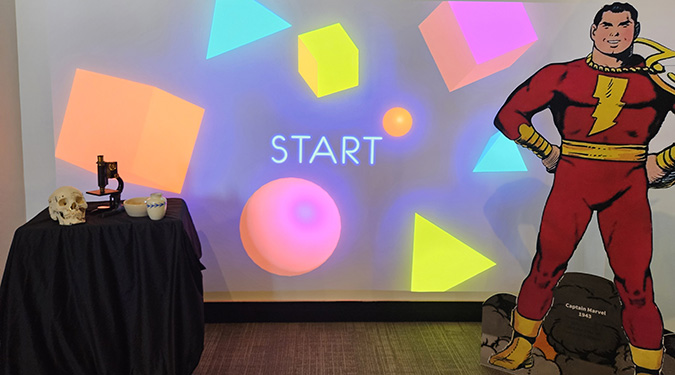 A colorful display features glowing geometric shapes with the word "START" in the center. A table with a skull and microscope is on the left, and a cartoon superhero cutout stands on the right. The scene mixes science and comic book themes.